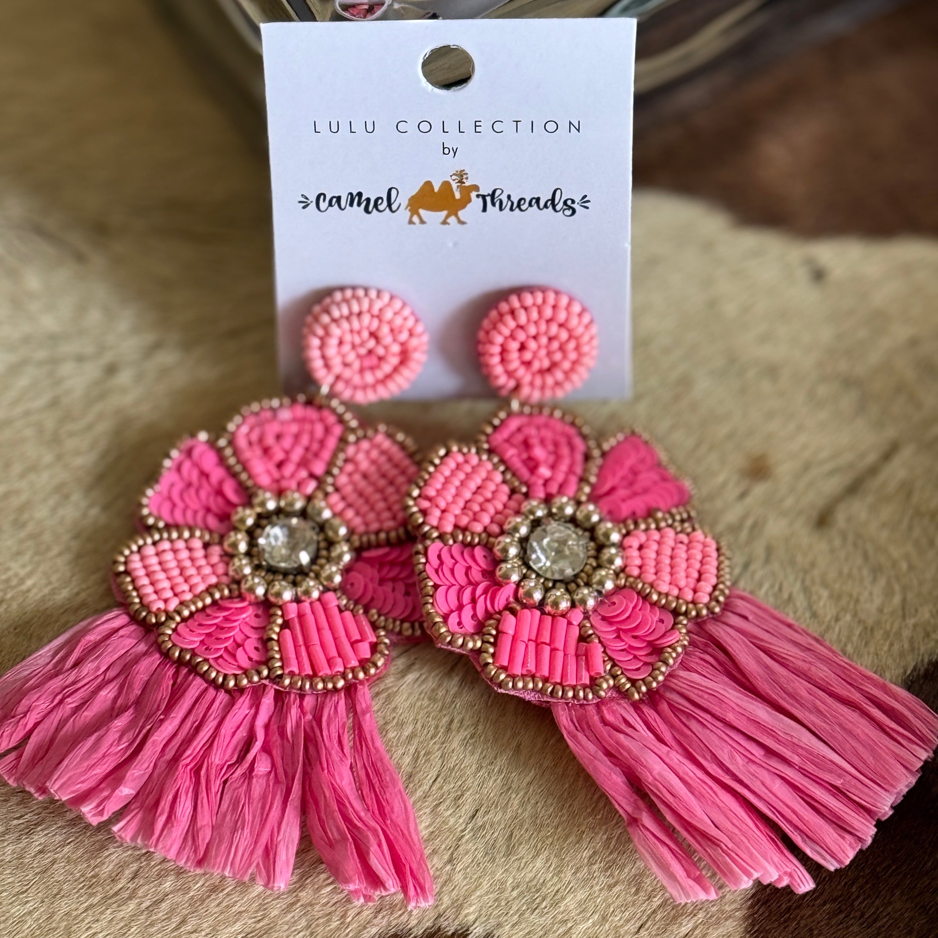A pair of pink beaded flower earrings with tassels, displayed on a white card with branding text.