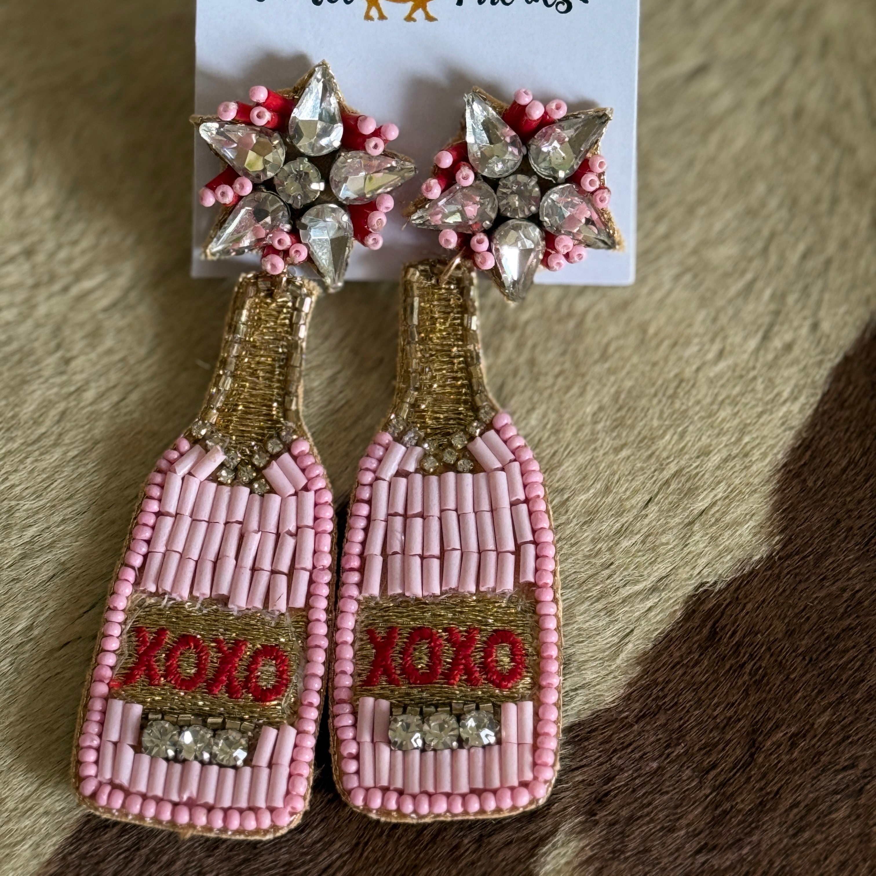 A pair of beaded earrings designed to resemble champagne bottles, with pink 'XOXO' text and gold details, displayed on a white card with 'LULU COLLECTION by Camel Threads' branding.