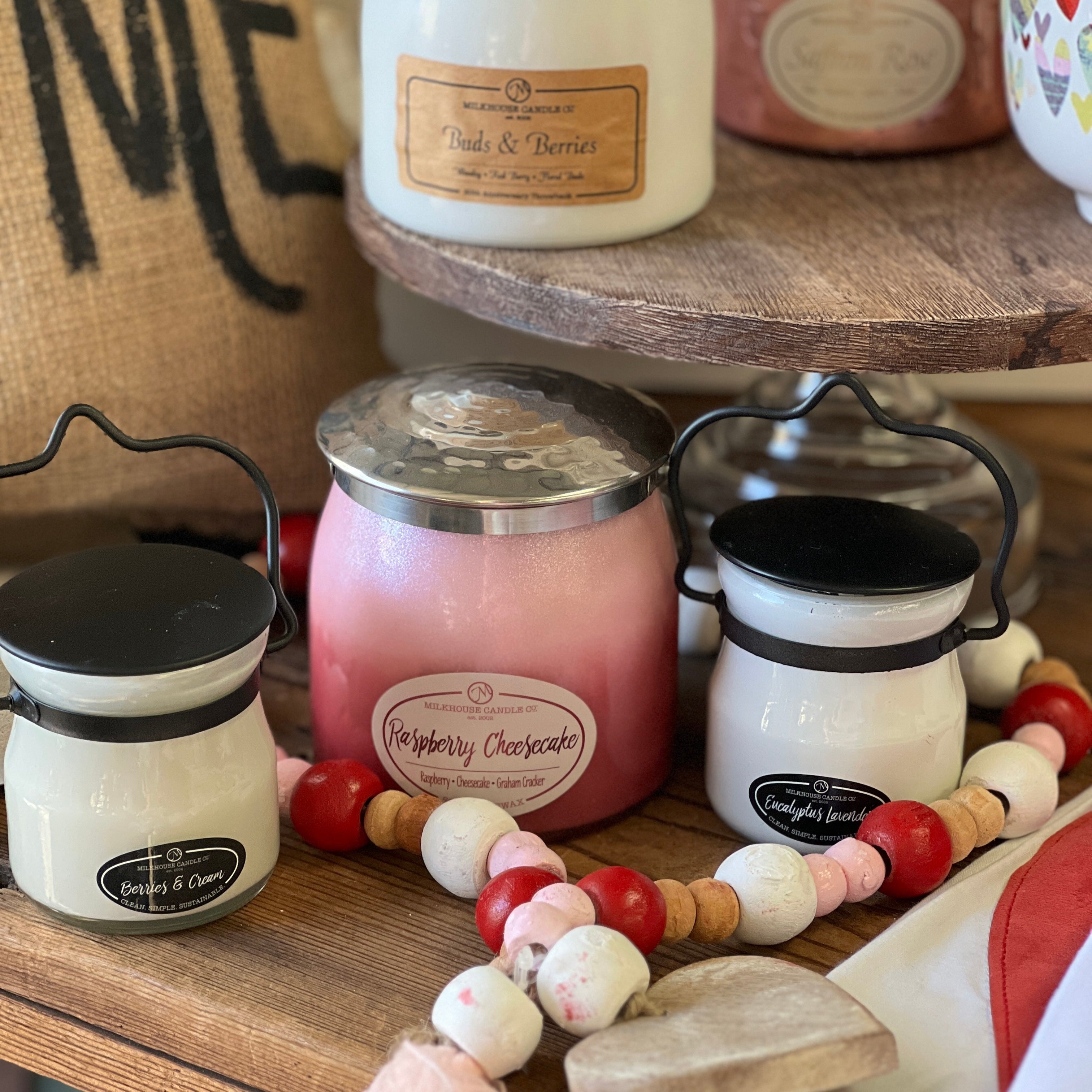 A set of Milkhouse brand candles, with one large jar and two smaller cream jars, displayed on a wooden surface with a 'Me' sign in the background. The candles have labels and the brand appears to be seasonal.