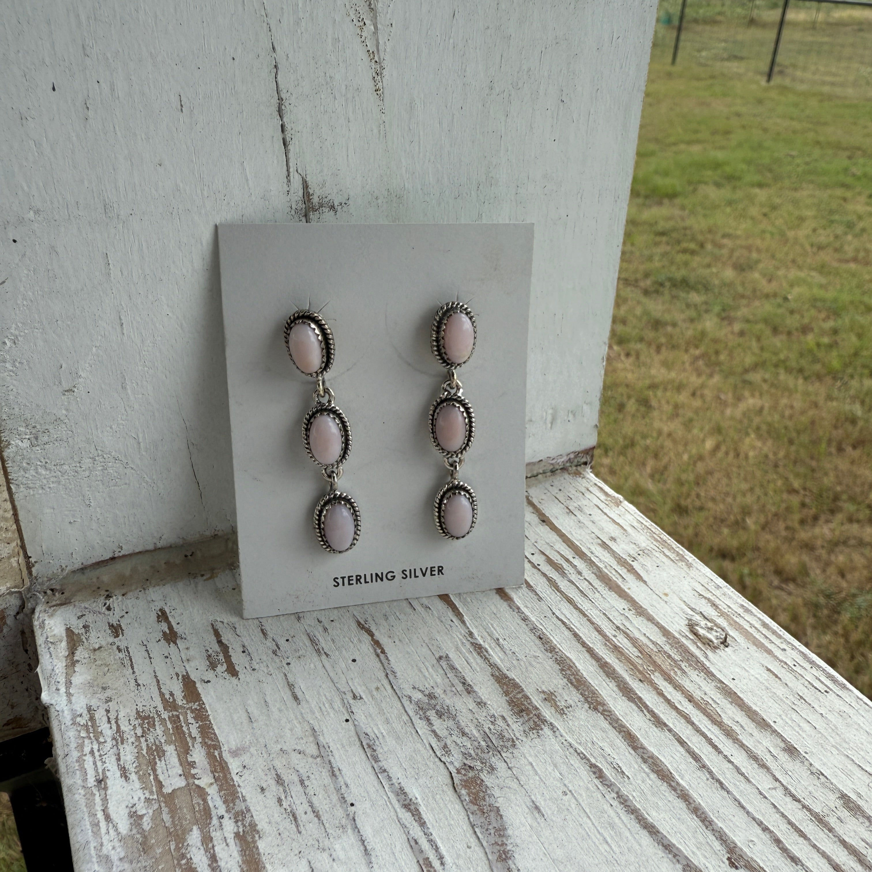 Pair of sterling silver pink opal earrings on a display card with a wooden surface and grass background