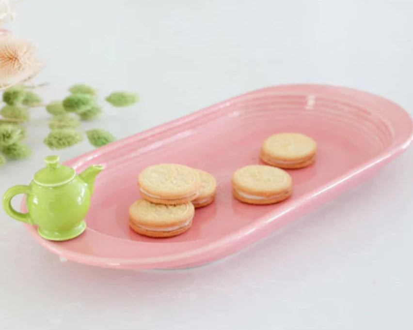 Pink ceramic tray with cookies and a green teapot on a white surface with flowers in the background.
