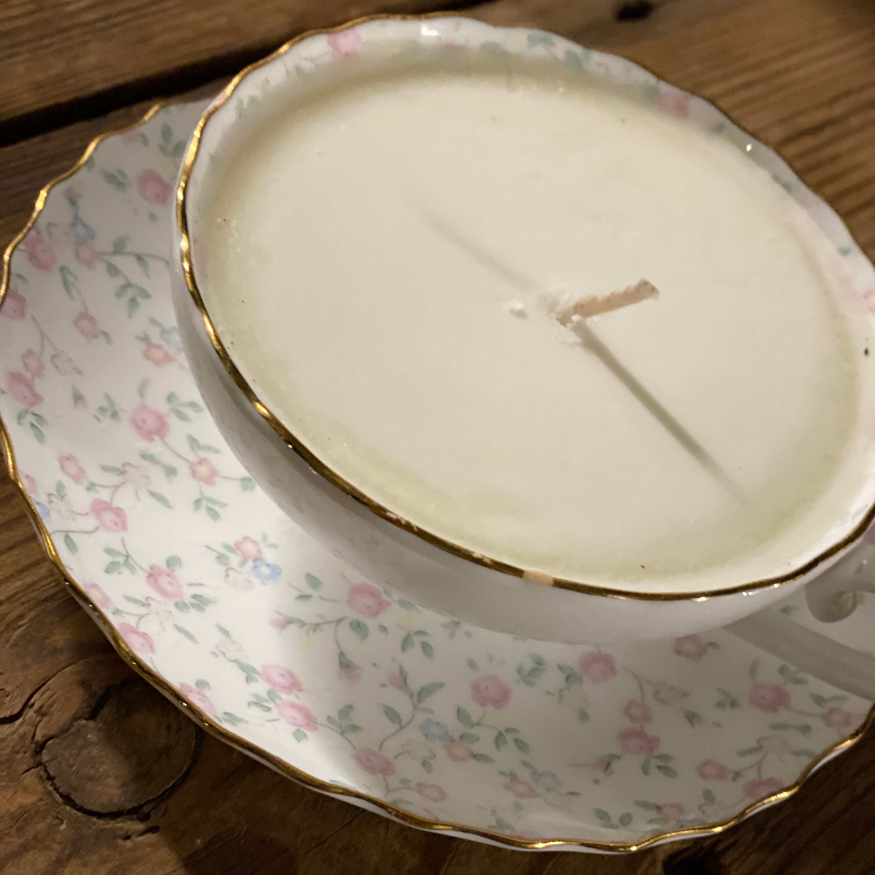A bone china vintage tea cup with gold trim and a floral pattern, featuring a candle inside, placed on a matching saucer.