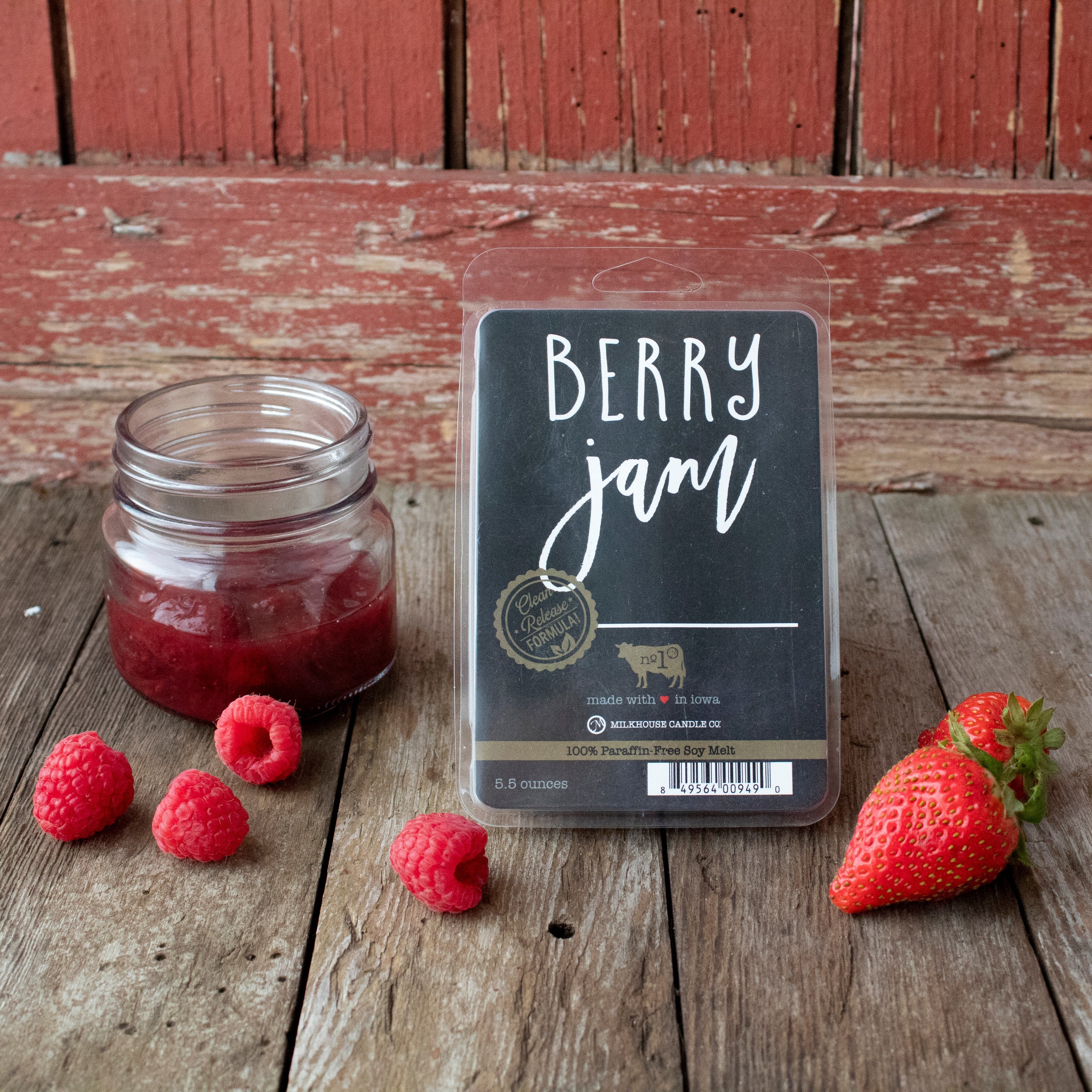 A wax melt pack labeled 'Berry Jam' is displayed next to a small clear jar with a red substance inside, and fresh strawberries and raspberries on a wooden surface.