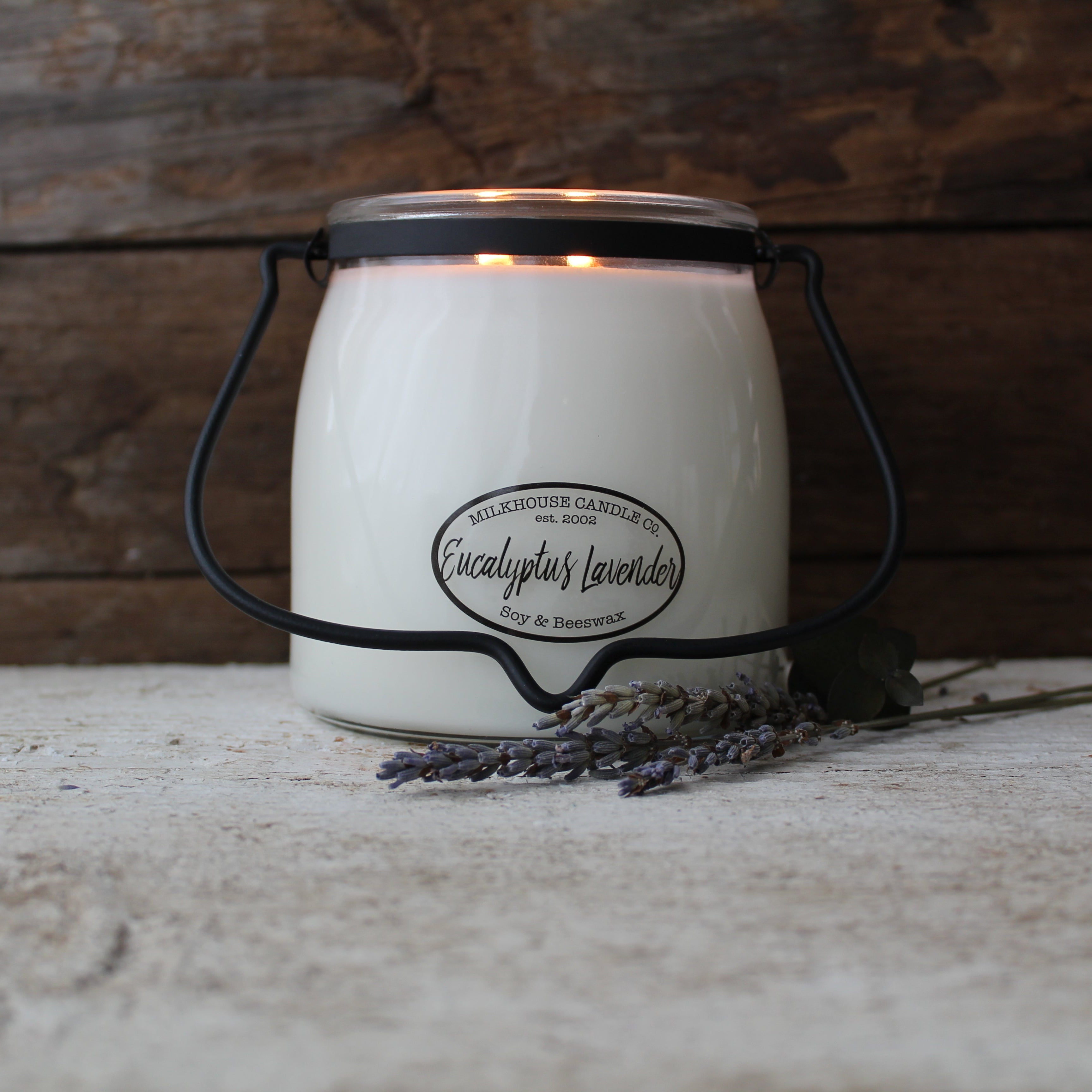 A white butter jar candle with black lid and label, set on a wooden surface with a sprig of lavender to the side.