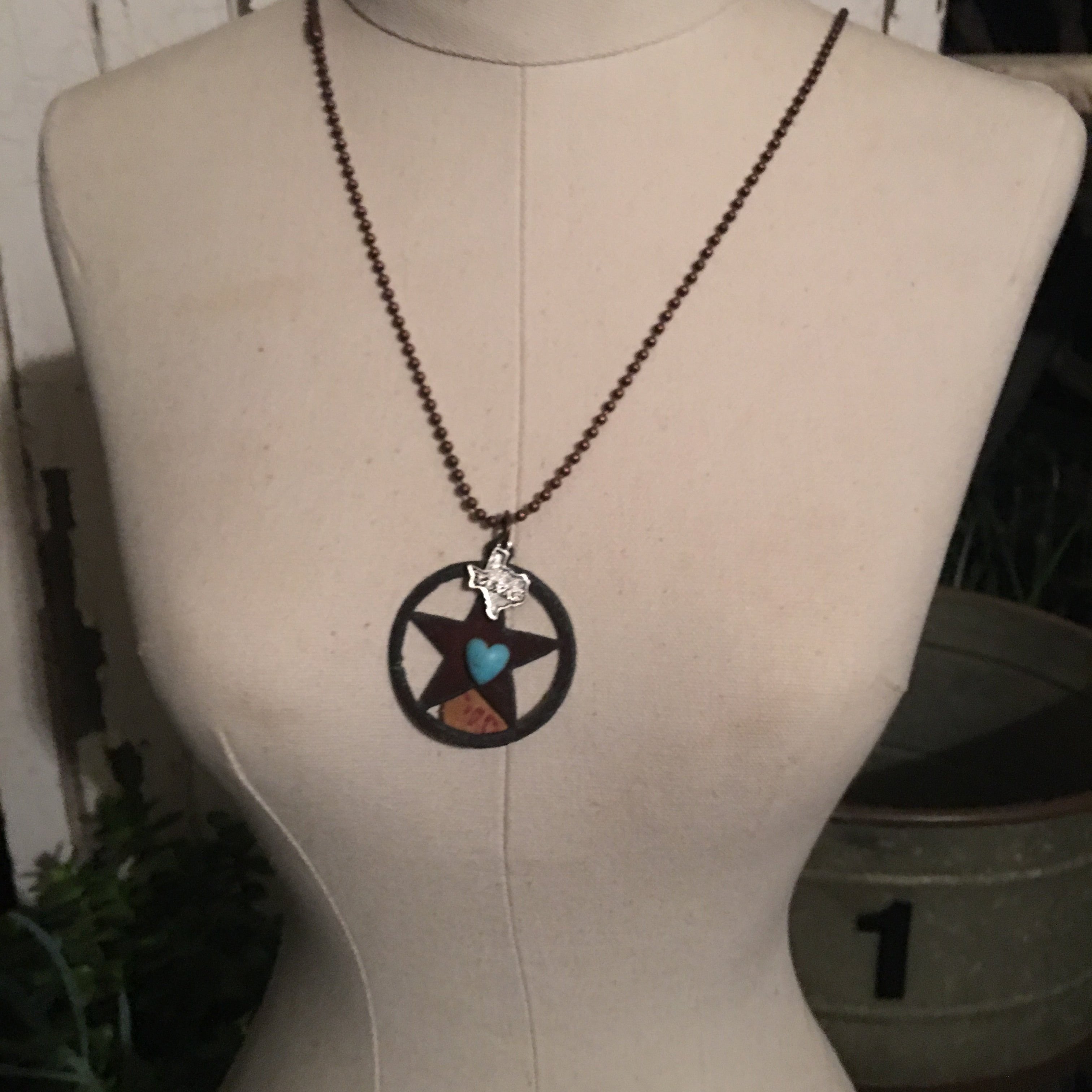 A necklace with rustic letters and shapes displayed on a mannequin. The letters appear to have a metal flower and a crystal center.