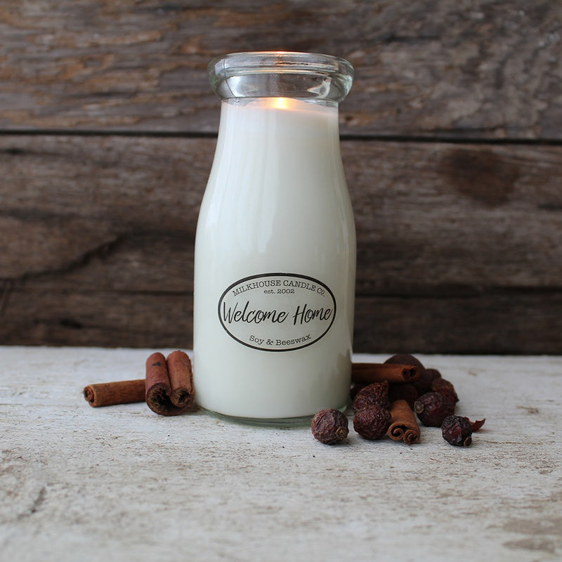 A white milk bottle candle with a label that reads 'Welcome Home', set on a wooden surface with some decorative elements like cinnamon sticks and pine cones around it.