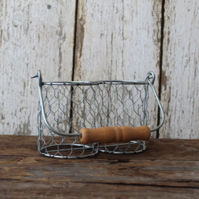 A silver metal basket with a rustic design, featuring a handle with a wooden grip, displayed on a wooden surface against a white wooden background.
