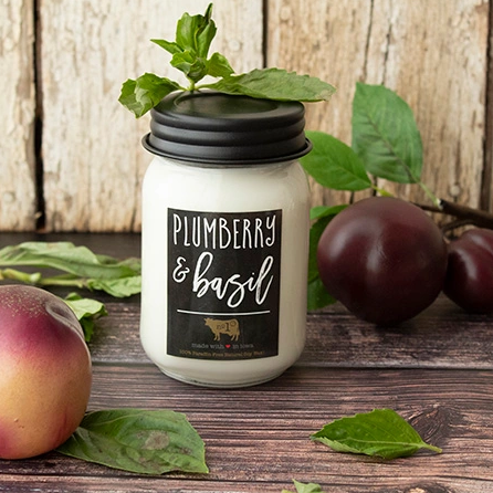 A Mason Jar Candle with a label that reads 'Plumberry & Basil'. The jar is white, and the candle is surrounded by fruits and leaves on a wooden background.